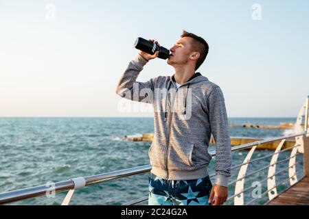 Profilo di bello sportivo in giacca e pantaloni corti bevendo acqua dalla bottiglia dopo la sessione di allenamento sul quay, vicino all'oceano. Sport concept. Foto Stock