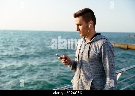 Bello pensieroso uomo in giacca sportiva e cuffie, guardando il display del cellulare, durante la deambulazione sul quay, vicino all'oceano. Foto Stock