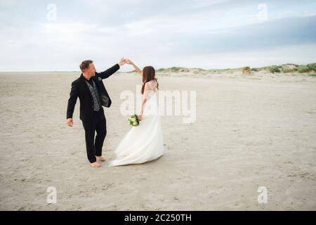 Gli sposi che ballano sulla spiaggia di sabbia. Sposa e sposo che si guardano l'uno all'altro e sorridono. Danza a piedi nudi. Concetto di giorno di nozze. Foto Stock