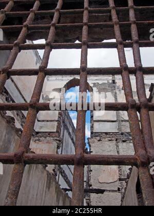 vista attraverso le ruste barre di ferro su una finestra di un edificio in rovina collassante con pareti sbriciolanti e aperto al cielo Foto Stock