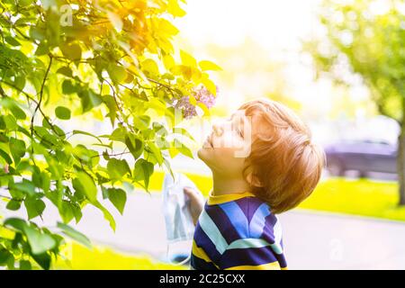 Ragazzo felice che togli la maschera medica protettiva e respirando aria, annusare fiori di lilla. Allegro bambino caucasico al sole giorno all'aperto nel parco Foto Stock