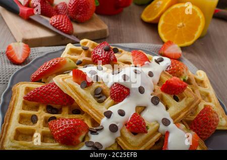 Waffles fatti in casa con lo sciroppo di acero e fragole, ricoperto di crema e scaglie di cioccolato, arancione bevanda fresca Foto Stock