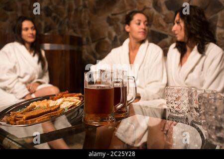 le giovani donne caucasiche felici in accappatoio sedersi accanto alla piscina, la sauna, bere birra fredda. Grazie Foto Stock