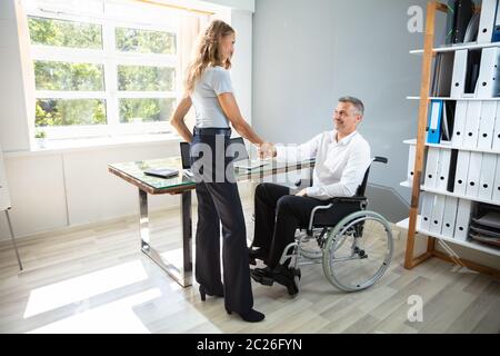 Happy Businessman disabili seduto su una sedia a rotelle agitando la mano con il suo partner in un ufficio moderno Foto Stock