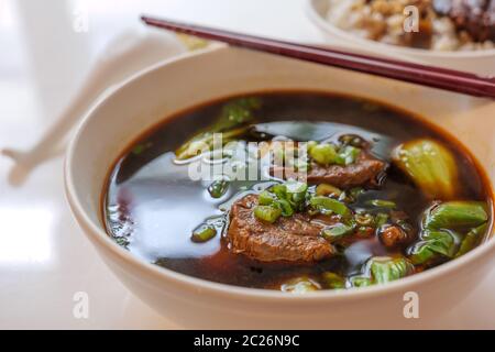 Vista del fuoco selezionata alla zuppa di noodle di manzo allo stufato taiwanese senza noodle in ciotola bianca sul tavolo bianco. Foto Stock