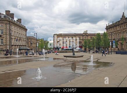 Persone che camminano in piazza st georges a Huddersfield tra le fontane d'acqua e gli edifici circostanti Foto Stock