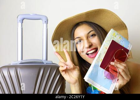 Ragazza ridente pronta per il viaggio mostrando segno di vittoria. Immagine di una donna viaggiatore pronta per i luoghi tropicali. Foto Stock