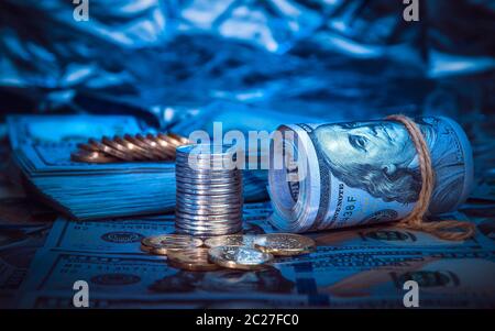 Un rotolo di dollari con le monete sullo sfondo di sparsi in un centinaio di fatture del dollaro nella luce blu Foto Stock