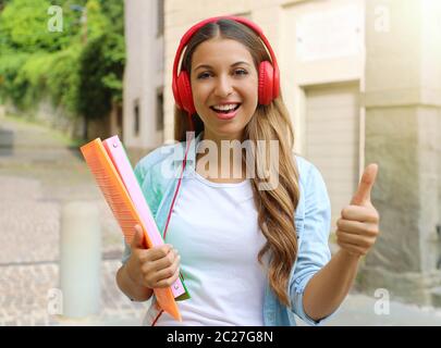 Felice ragazza dello studente con le cuffie dà il pollice fino all'esterno. Giovane donna facendo un programma di scambio studentesco. Foto Stock