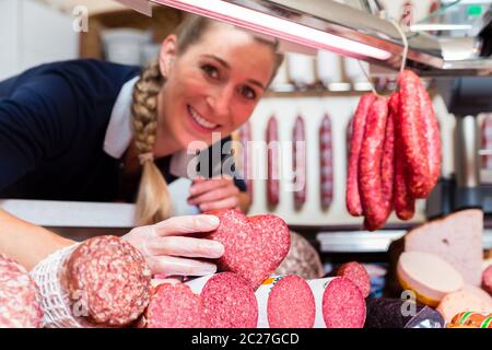 Donna di vendite nel negozio di carne che mostra un cuore salsiccia sagomato per il cliente Foto Stock