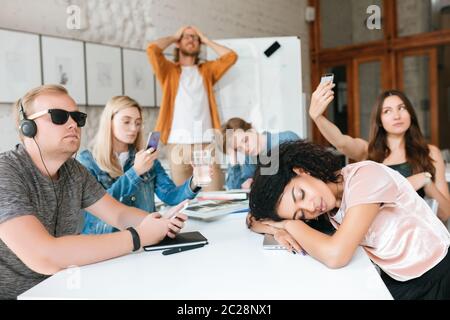 Insegnante stanco in piedi vicino a bordo e tenendo le mani sulla testa. Gruppo di giovani studenti che fanno foto, ascoltano musica e dormono durante la lezione. STU Foto Stock