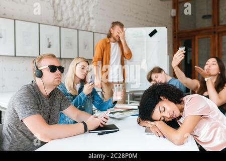 Giovane insegnante stanco in piedi vicino a bordo e coperto il suo viso con la mano. Gruppo di giovani studenti che fanno foto, ascoltano musica e dormono di meno Foto Stock