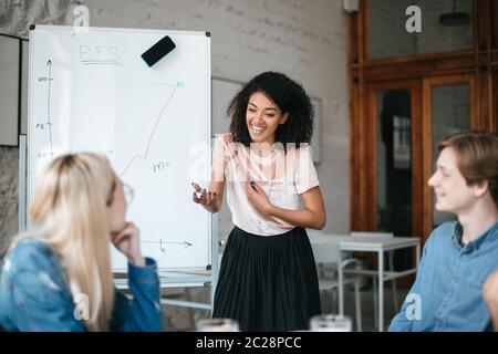 Ritratto di felice afroamericana ragazza in piedi vicino a bordo e discutere di nuovo progetto con i colleghi in ufficio . Gruppo di persone sorridenti che si siedono a. Foto Stock