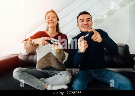 Divertente coppia giovane i giochi per computer con joystick, seduto sul lettino, a casa. Vestito di maglioni e jeans. Foto Stock