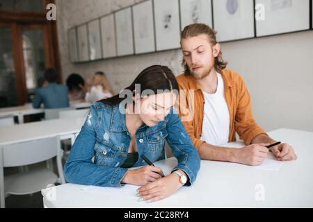 Ritratto del giovane che salpava alla prova del compagno di classe. Serio ragazza che scrive il test pensieroso mentre lo chiude a mano. Due studenti seduti al Foto Stock