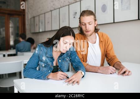 Giovane uomo che salpava con cura nella prova del compagno di classe in lezione. Seria ragazza che scrive testando pensieroso e chiudendolo a mano. Due studenti seduti Foto Stock