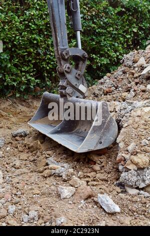 Scavo di un concreto passo carraio al fianco di una siepe; metallo benna di scavo si appoggia su un gran mucchio di rotture di calcestruzzo e mattoni Foto Stock