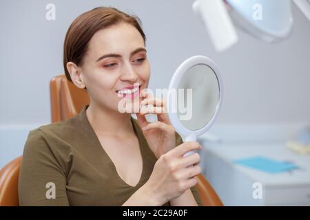 Giovane donna attraente sorridente, esaminando i denti nello specchio, mentre visita dentista. Donna che gode di risultati professionali di pulizia dei denti e wh Foto Stock