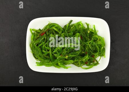 Primo piano di insalata verde di alghe wakame su piatto bianco su sfondo nero della tavola di ardesia, vista dall'alto, direttamente sopra Foto Stock