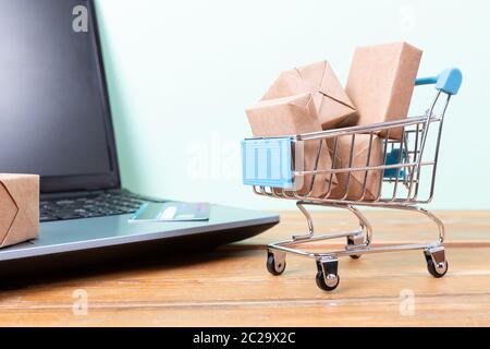 Shopping online concetto. Carrello spesa, piccole scatole, laptop e una carta di credito sulla scrivania Foto Stock