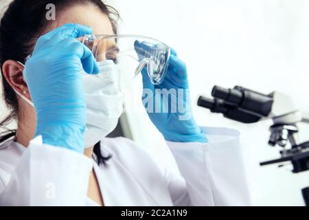 Donna scienziato mette su occhiali protettivi. Giovane donna medico in guanti medici e maschera viso mette su occhiali protettivi con microscopio nel backg Foto Stock