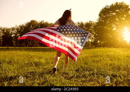 Vista posteriore di giovane donna con bandiera americana che corre verso il tramonto in campagna. Festa del giorno dell'indipendenza Foto Stock