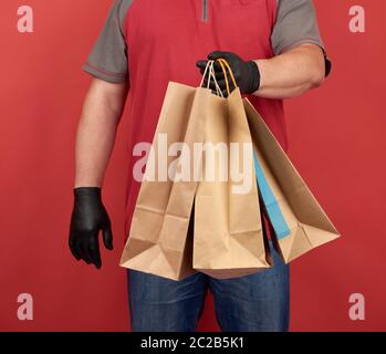 Uomo in una T-shirt rossa, con guanti in lattice nero, contiene sacchetti di carta per l'acquisto, consegna sicura degli ordini, sfondo corax Foto Stock