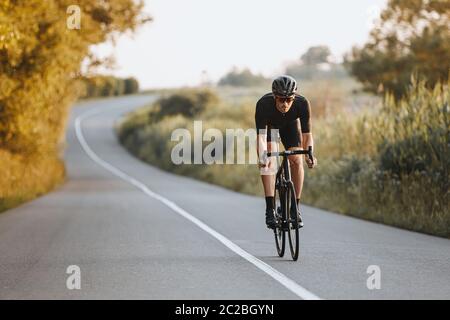 Ciclista maschile professionale in casco nero, occhiali protettivi e abbigliamento sportivo in bicicletta dinamica su strada asfaltata con sfondo sfocato. Concetto di Foto Stock