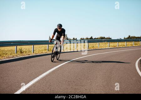 Uomo maturo attivo in outfit sportivo in bicicletta su strada asfaltata in campagna. Ciclista esperto in casco nero e occhiali a specchio regolarmente trai Foto Stock