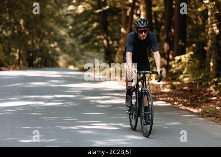 Ciclista professionale in abbigliamento sportivo e casco nero in bicicletta su strada asfaltata tra foresta estiva. Atleta portandalchi in occhiali protettivi traini Foto Stock