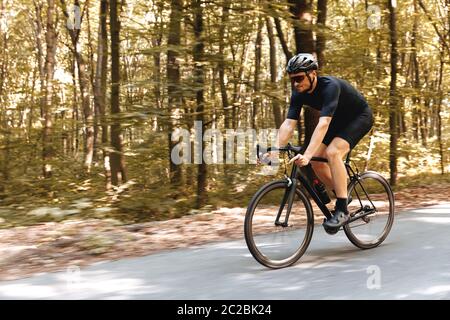 Ciclista bearded in sportswear e vetri specchiati che guidano dinamicamente la bicicletta su strada asfaltata. Sportivo professionista in casco protettivo godendo favo Foto Stock
