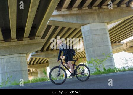 Il ciclista che guida una bicicletta su una strada aperta per l'atleta al tramonto, il ciclista di ragazzo bearded, corre sull'autostrada su una bici sportiva. Sotto il ponte. All'aperto. S Foto Stock