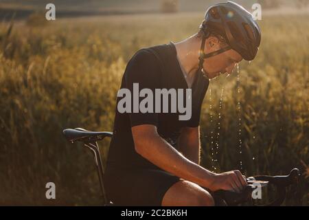 Vista laterale del ciclista maturo con viso umido stanco che indossa casco nero e abbigliamento sportivo seduto in bicicletta all'aperto. Uomo sopportato che prende un af di riposo Foto Stock