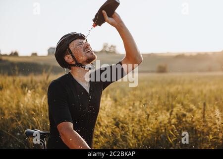 Atleta maturo stanco in abbigliamento sportivo e casco protettivo che spruzzi acqua fredda sul viso dalla bottiglia nera dopo aver cavalcato in bicicletta durante le giornate di sole. Barba Foto Stock