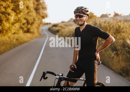 Uomo allegro e portatore in abbigliamento attivo, casco nero e occhiali sportivi seduti in bicicletta e guardando la fotocamera con bella natura intorno. Concetto di acti Foto Stock