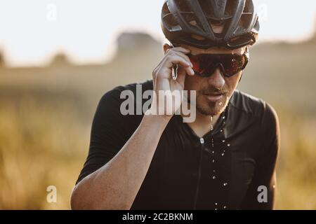 Ritratto di uomo bearded in casco nero e occhiali specchiati sensazione stanco a causa di duro allenamento. Ciclista matura con viso bagnato che prende la pausa durante Foto Stock