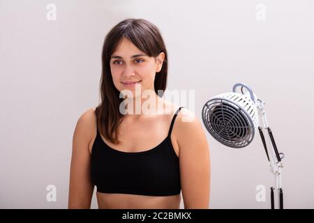 Sorridente giovane donna riceve la radiazione infrarossa di calore lampada luce terapia contro lo sfondo grigio Foto Stock