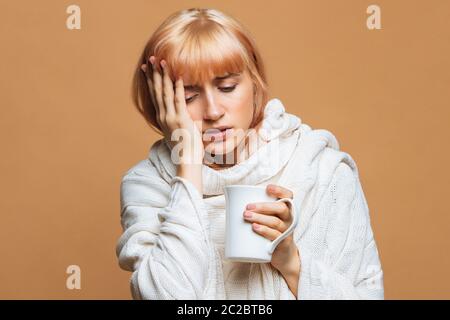 Malattia giovane donna carina con capelli biondi fragola, plaid caldo con mal di testa, tenendo la tazza di tè caldo, toccando la fronte, sentire il primo symp Foto Stock