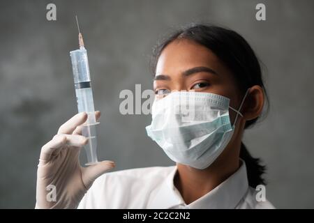 Primo piano di una giovane dottoressa con siringa. Donna medico in maschera protettiva ritratto su fondo parete in cemento Foto Stock