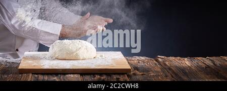 Chef o baker spolvero un mucchio di pasta cruda con farina sospesa in aria battendo le mani in un panorama rustico banner con spazio di copia Foto Stock