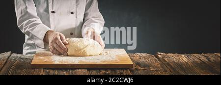 Il cuoco in una pizzeria o baker preparare un tumulo di pasta cruda su una infarinata scheda di legno su un tavolo rustico in un panorama banner con spazio di copia Foto Stock