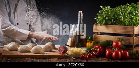 Lo Chef battendo la sua mani infarinata creando una nuvola di farina in aria come egli prepara le porzioni di pasta per pizza su una tabella con fresca ingredien assortiti Foto Stock