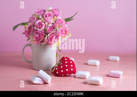 Di piccole dimensioni e di colore bianco e rosa marshmallows sono sparsi su un pallido sfondo rosa accanto a un vaso di rose. Posto per il testo. Foto Stock