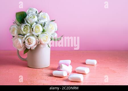 Di piccole dimensioni e di colore bianco e rosa marshmallows sono sparsi su un pallido sfondo rosa accanto a un vaso di rose. Posto per il testo. Foto Stock