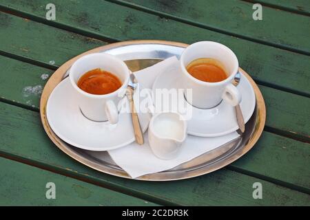 Chiudere uno argento vassoio di metallo bianco con tazze di caffè espresso su piattini con cucchiai e bricco per latte servita sul verde di legno verniciato tabella, elevato h Foto Stock