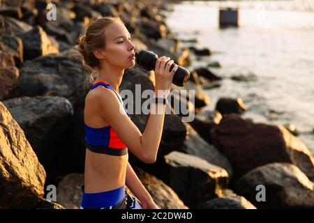 Attraente ragazza pensosa in sportswear, bevendo acqua dalla bottiglia, seduti su pietre di mare. Un sano concetto. Foto Stock