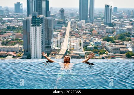 Un giovane uomo felice si rilassa sul bordo della piscina sul tetto dell'hotel Foto Stock