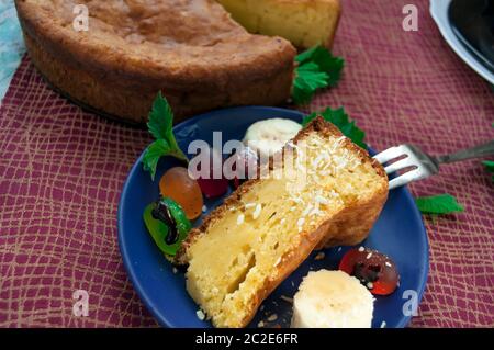 Torta su una tovaglia di colore bordeaux. Un pezzo di torta con caramelle di gelatina, fette di banana e fiocchi di cocco è preso con una forchetta su un piatto blu Foto Stock