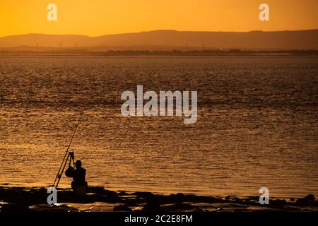 Silhouette di un uomo che pesca nell'estuario del Severn al largo della costa di Portishead al tramonto con la costa gallese in lontananza. Foto Stock