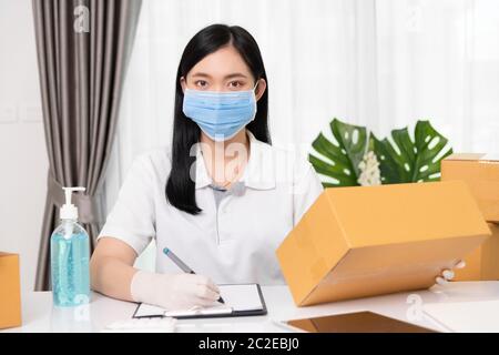 Giovane asiatico piccolo imprenditore di affari che indossa maschera di protezione per trasportare le scatole del prodotto all'ufficio domestico, imprenditore di PMI di avvio o donna freelance WO Foto Stock